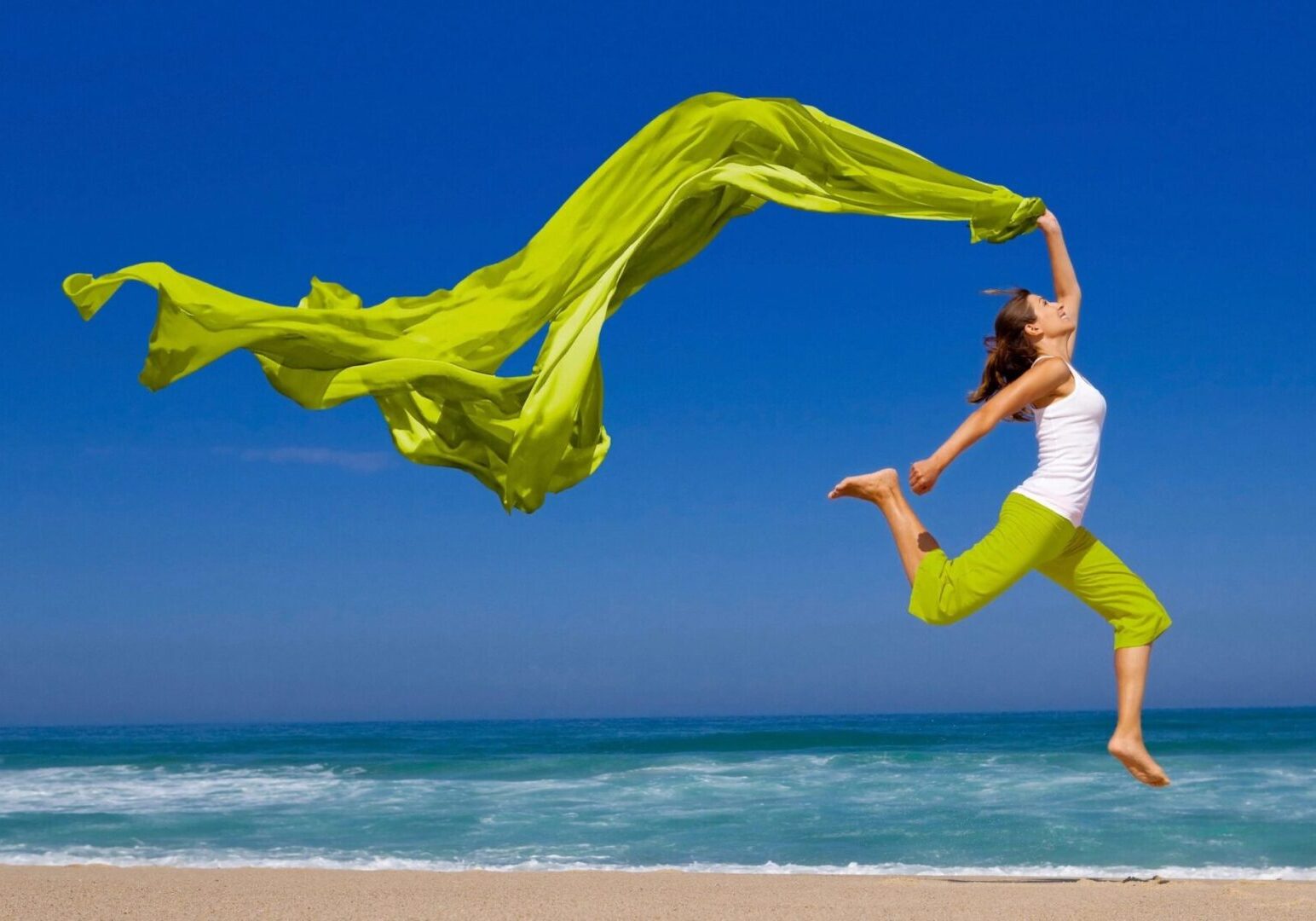 A woman jumping on the beach with a green scarf.